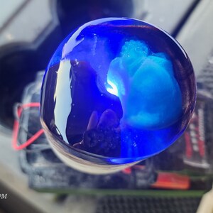 May include: A close-up of a blue glass sphere with a swirling blue and white pattern inside. The sphere is sitting on a white surface.