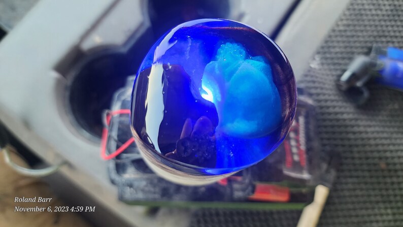 May include: A close-up of a blue glass sphere with a swirling blue and white pattern inside. The sphere is sitting on a white surface.