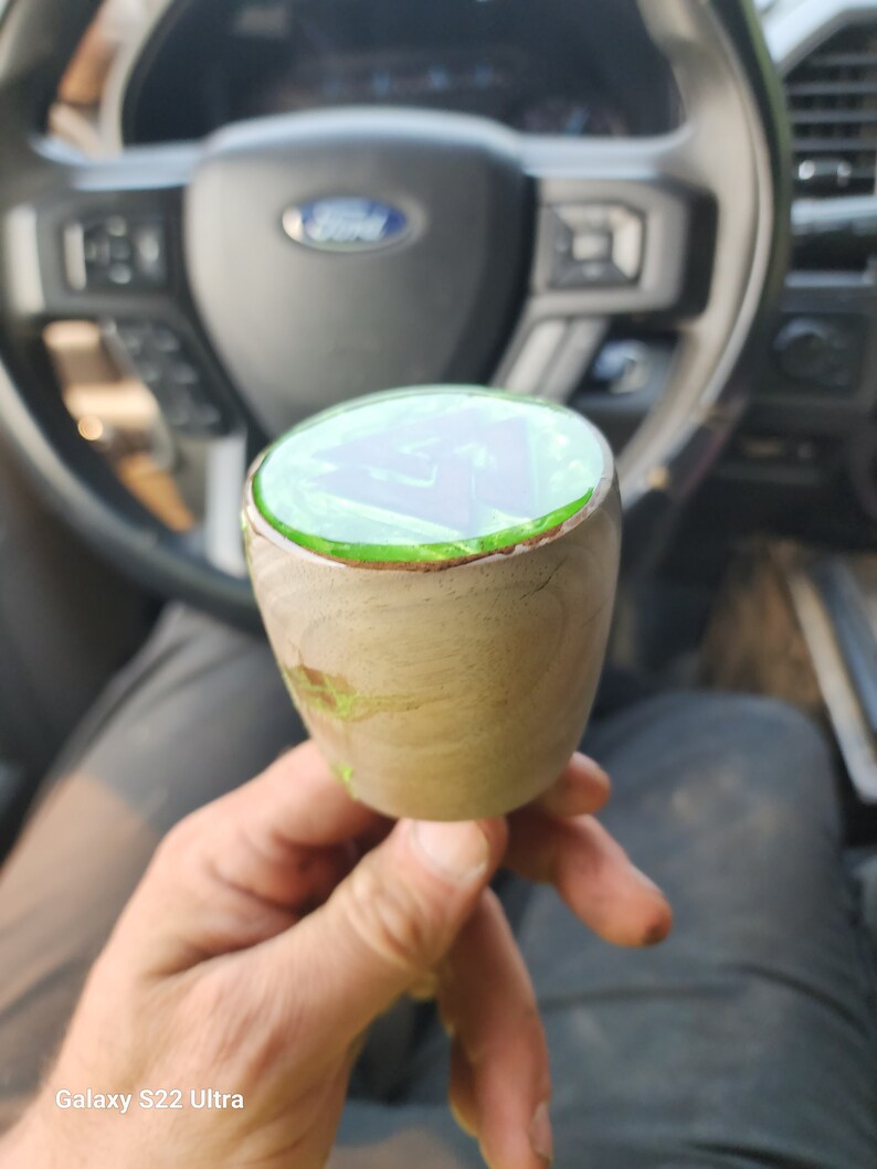 May include: A close-up of a wooden gear shift knob with a green epoxy resin top. The resin has a swirl pattern and is clear in the centre.