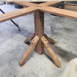 May include: A wooden table base with four legs and a square top. The base is made of dark brown wood with a natural grain pattern. The table base is being assembled with a clamp.