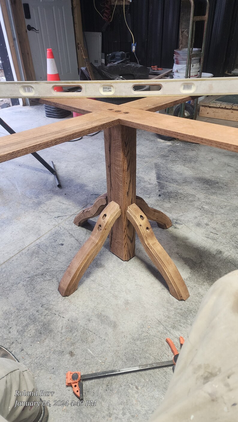 May include: A wooden table base with four legs and a square top. The base is made of dark brown wood with a natural grain pattern. The table base is being assembled with a clamp.