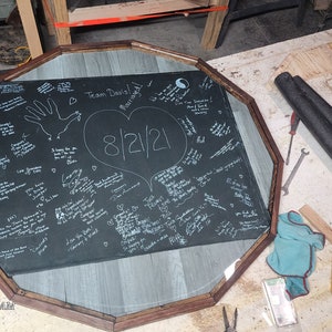 May include: An octagonal sign with a black background and white writing, framed in wood. The sign features a large heart with the date "8/12/21" inside. The text "Team Davis" is written at the top of the sign.