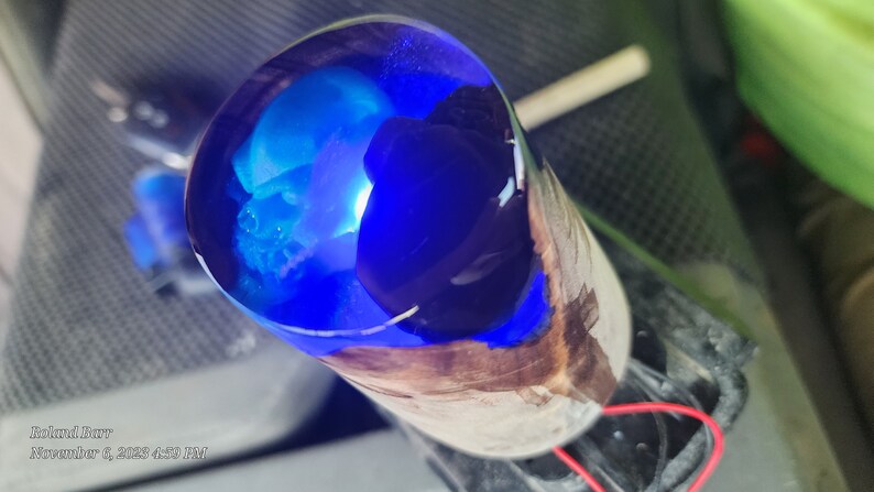 May include: A close-up of a blue and black resin cast inside a wooden cylinder. The resin is clear and has a blue and black swirl pattern. The wood is brown and has a natural grain.