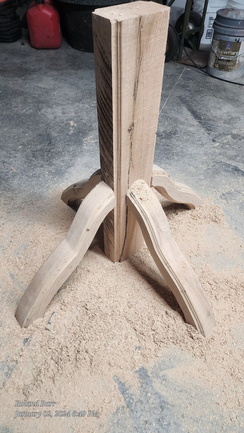May include: A wooden table base with four legs that are curved and joined at the top. The base is made of unfinished wood and is sitting on a pile of sawdust.
