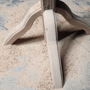May include: Close-up of a wooden table base with three legs. The legs are curved and have a decorative design. The base is surrounded by sawdust.