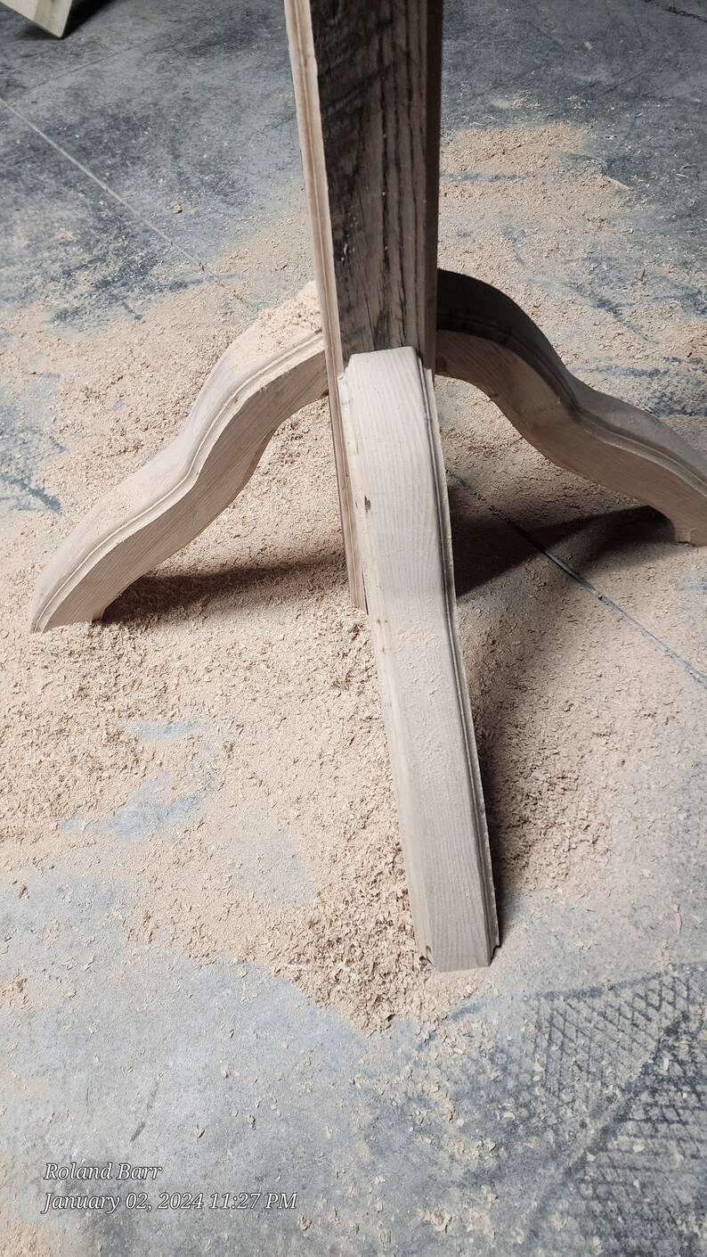 May include: Close-up of a wooden table base with three legs. The legs are curved and have a decorative design. The base is surrounded by sawdust.