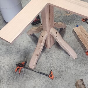 May include: A wooden table base with four legs and a square top. The base is made of natural wood with a light brown finish. The table top is made of a lighter wood and is attached to the base with screws. The table is being assembled on a concrete floor.
