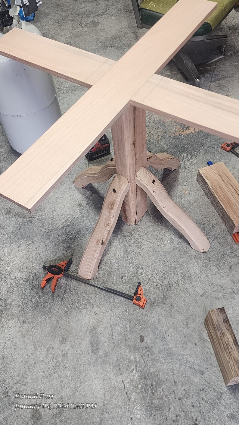 May include: A wooden table base with four legs and a square top. The base is made of natural wood with a light brown finish. The table top is made of a lighter wood and is attached to the base with screws. The table is being assembled on a concrete floor.