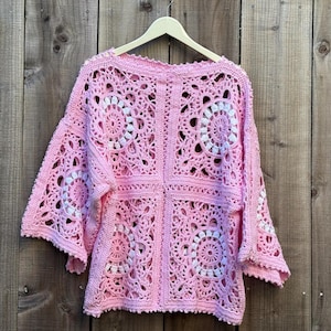 May include: A pink crochet top with white circular patterns, displayed on a wooden hanger. The openwork design features square panels with floral-like motifs. The top is made of soft yarn and has wide sleeves.