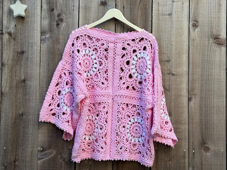 May include: A pink crochet top with white circular patterns, displayed on a wooden hanger. The openwork design features square panels with floral-like motifs. The top is made of soft yarn and has wide sleeves.