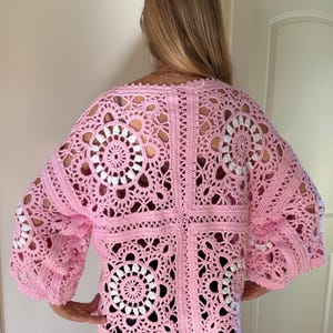 May include: A light pink crochet top with a square pattern and openwork design. The top features a floral motif in white and pink, with wide sleeves. The back view shows the intricate details of the handmade garment.