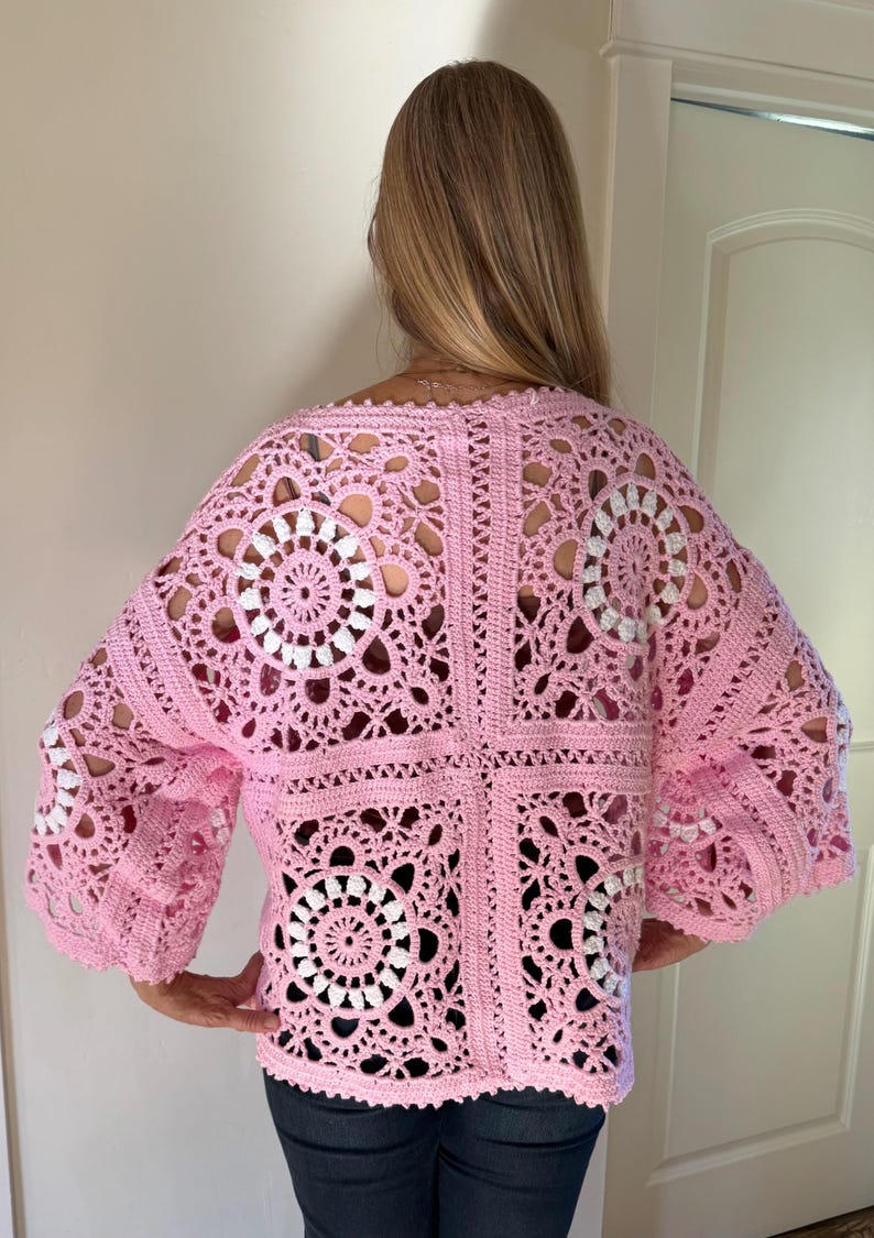 May include: A light pink crochet top with a square pattern and openwork design. The top features a floral motif in white and pink, with wide sleeves. The back view shows the intricate details of the handmade garment.