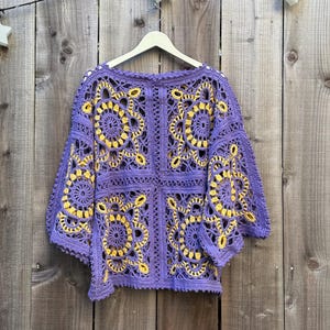 May include: A purple crochet top with yellow floral patterns. The top is made of a soft, open-weave material and has a round neckline and short sleeves. The top is hanging on a white hanger against a wooden background.