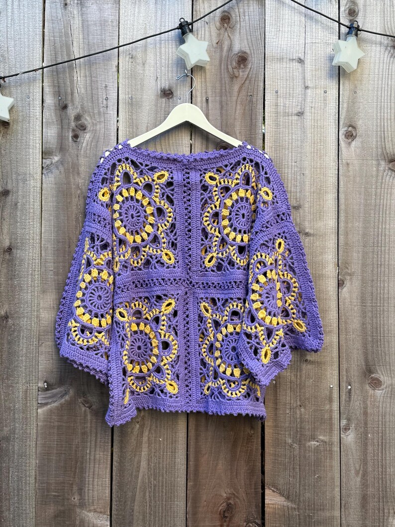 May include: A purple crochet top with yellow floral patterns. The top is made of a soft, open-weave material and has a round neckline and short sleeves. The top is hanging on a white hanger against a wooden background.