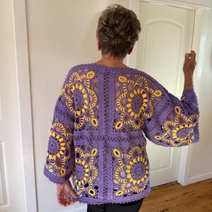 May include: A purple crochet jacket with yellow floral patterns. The jacket features a loose, open design with wide sleeves. The crochet work is detailed, showcasing the intricate floral motifs. The jacket is a handmade garment.
