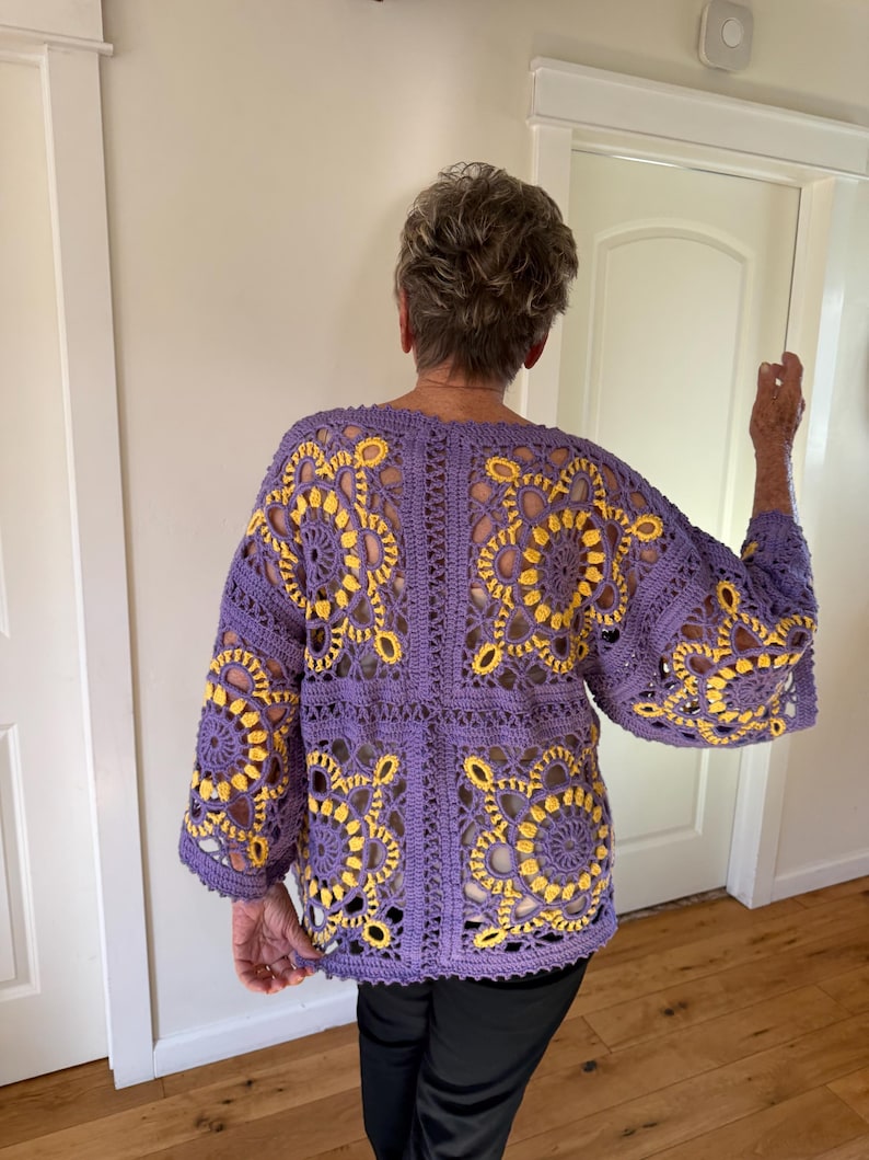 May include: A purple crochet jacket with yellow floral patterns. The jacket features a loose, open design with wide sleeves. The crochet work is detailed, showcasing the intricate floral motifs. The jacket is a handmade garment.