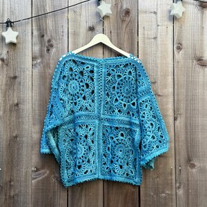 May include: A turquoise crochet top with a square pattern and long sleeves hangs on a wooden hanger. The openwork design reveals the texture of the yarn. The background is a wooden fence with star-shaped lights.