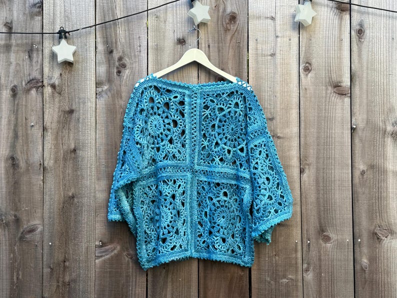 May include: A turquoise crochet top with a square pattern and long sleeves hangs on a wooden hanger. The openwork design reveals the texture of the yarn. The background is a wooden fence with star-shaped lights.