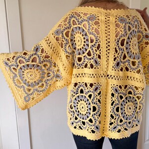 May include: A yellow crochet top with gray circular and floral patterns. The open-weave design features wide sleeves and a square body. The garment is handmade and appears to be worn over dark pants.