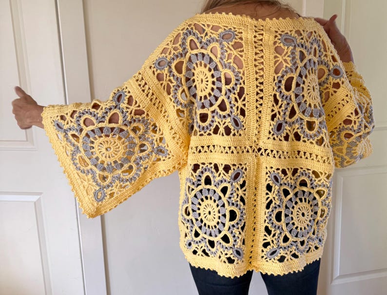 May include: A yellow crochet top with gray circular and floral patterns. The open-weave design features wide sleeves and a square body. The garment is handmade and appears to be worn over dark pants.