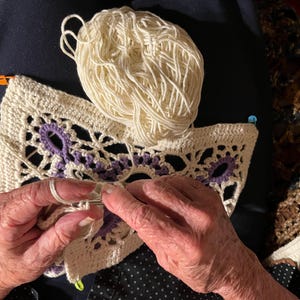 May include: Cream-colored crochet project with purple accents and a ball of yarn. The crochet work features an open, lacy design. The image shows the hands of a person working on the crochet project with a crochet hook.