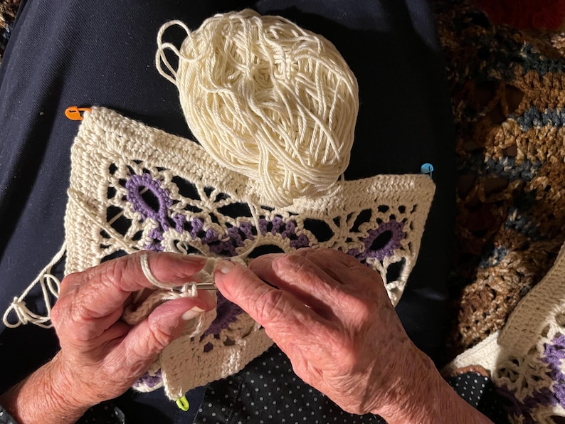 May include: Cream-colored crochet project with purple accents and a ball of yarn. The crochet work features an open, lacy design. The image shows the hands of a person working on the crochet project with a crochet hook.
