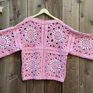 May include: A light pink, open-front crochet cardigan with a square pattern. The design features white circular accents within the squares. The cardigan is displayed on a wooden hanger against a wooden background.