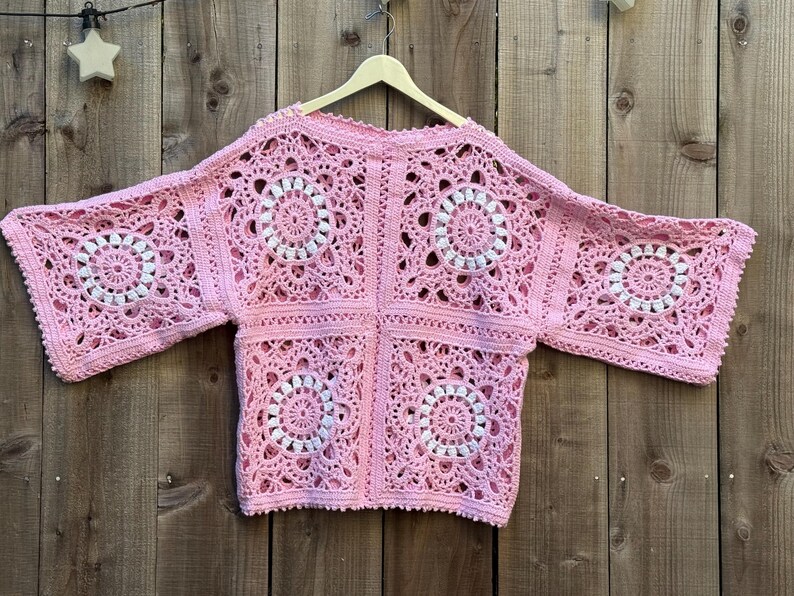 May include: A light pink, open-front crochet cardigan with a square pattern. The design features white circular accents within the squares. The cardigan is displayed on a wooden hanger against a wooden background.