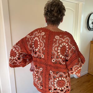 May include: A rust-colored, open-weave crochet top with a floral design. The top features a square pattern with white accents, creating a striking contrast. The sleeves are wide and the overall style is loose and comfortable.