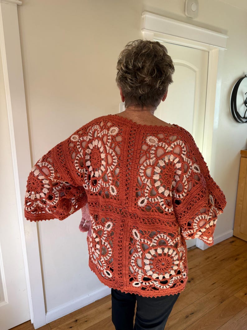 May include: A rust-colored, open-weave crochet top with a floral design. The top features a square pattern with white accents, creating a striking contrast. The sleeves are wide and the overall style is loose and comfortable.
