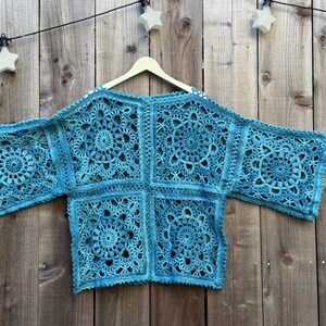 May include: A turquoise crochet top with a floral design, hanging on a wooden hanger. The openwork garment is made of squares with circular patterns. The background is a wooden fence.
