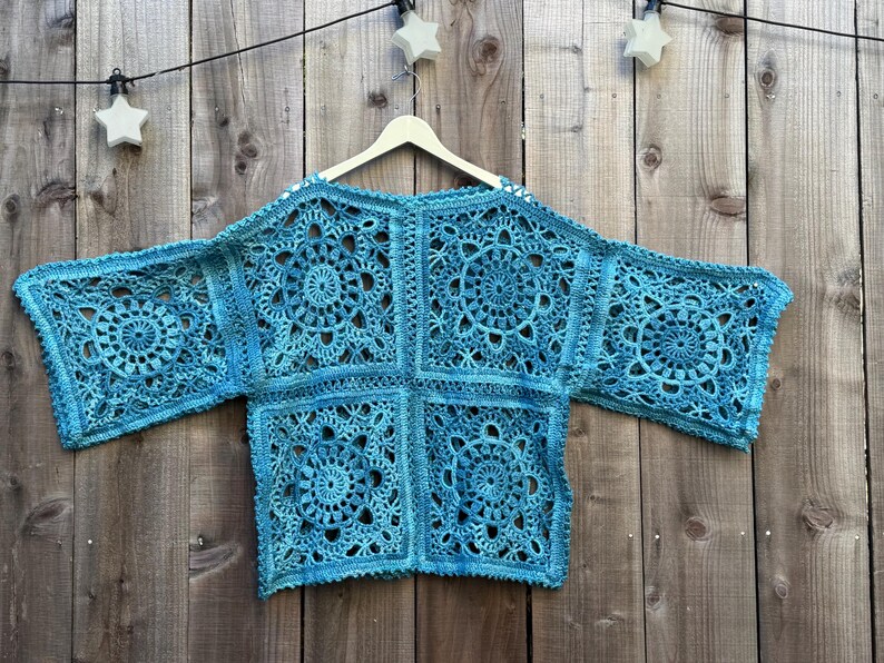 May include: A turquoise crochet top with a floral design, hanging on a wooden hanger. The openwork garment is made of squares with circular patterns. The background is a wooden fence.