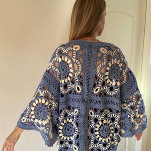 May include: A blue crochet cardigan with a floral design. The open-weave pattern features cream-colored accents, creating a contrasting visual texture. The cardigan has wide sleeves and a relaxed fit, perfect for layering.