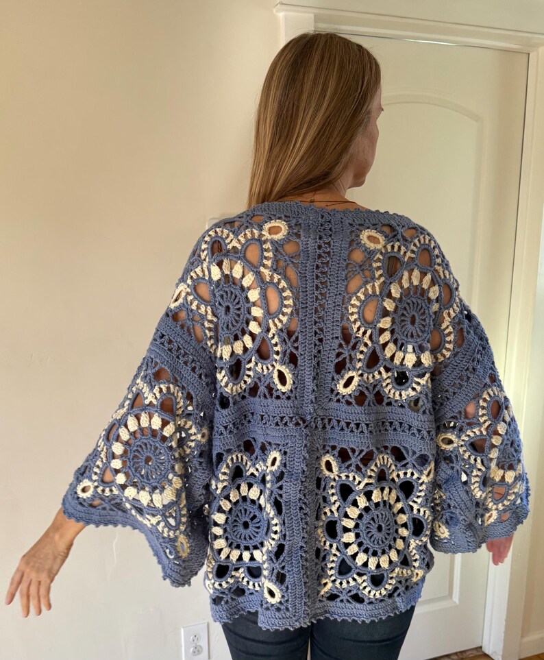 May include: A blue crochet cardigan with a floral design. The open-weave pattern features cream-colored accents, creating a contrasting visual texture. The cardigan has wide sleeves and a relaxed fit, perfect for layering.