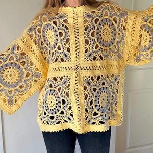 May include: A yellow crochet top with a square pattern design. The top features a floral design in gray and yellow. The sleeves are wide and flared. The top is made of a soft, open-weave material.