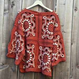 May include: Rust-colored crochet top with white floral designs. The openwork sweater is hanging on a wooden hanger against a wooden background. The sleeves are wide and bell-shaped, and the top is made of a soft, textured yarn.