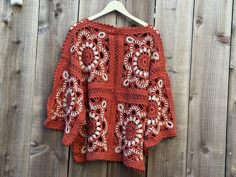 May include: Rust-colored crochet top with white floral designs. The openwork sweater is hanging on a wooden hanger against a wooden background. The sleeves are wide and bell-shaped, and the top is made of a soft, textured yarn.