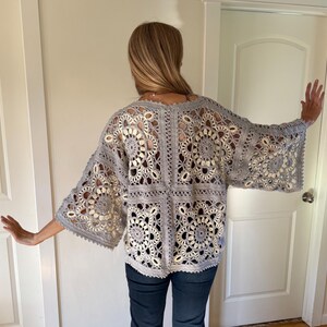 May include: A gray, open-weave crochet top with bell sleeves. The top features a floral pattern and is worn over a light-colored garment. The crochet design is visible, creating an airy and textured look. The top is paired with dark blue jeans.