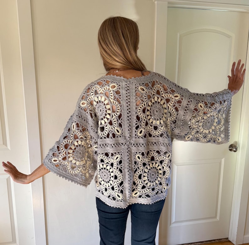 May include: A gray, open-weave crochet top with bell sleeves. The top features a floral pattern and is worn over a light-colored garment. The crochet design is visible, creating an airy and textured look. The top is paired with dark blue jeans.