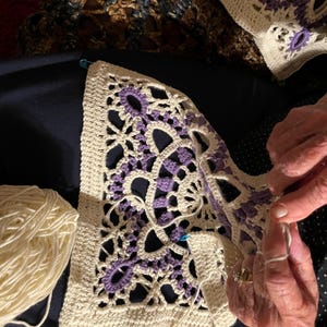 May include: Close-up of a crochet project in progress. The piece features a cream-colored background with intricate purple floral designs. A ball of cream-colored yarn sits nearby. The crafter's hands are visible, working with a crochet hook and yarn.