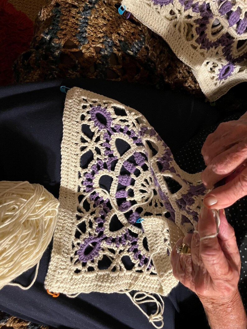 May include: Close-up of a crochet project in progress. The piece features a cream-colored background with intricate purple floral designs. A ball of cream-colored yarn sits nearby. The crafter's hands are visible, working with a crochet hook and yarn.