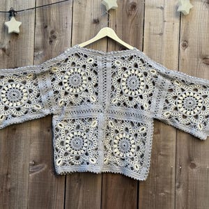 May include: A light gray, open-front crochet top with a floral pattern. The top is displayed on a wooden hanger against a wooden background. The sleeves are wide and the design is intricate.