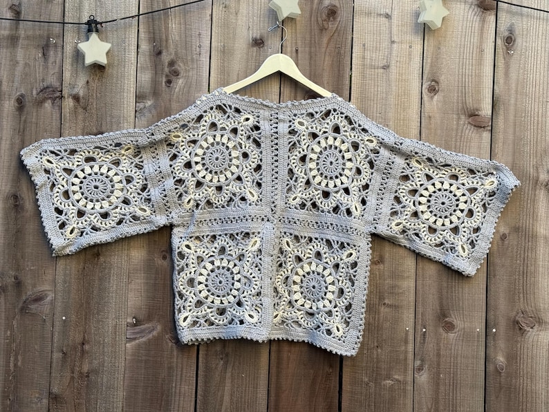 May include: A light gray, open-front crochet top with a floral pattern. The top is displayed on a wooden hanger against a wooden background. The sleeves are wide and the design is intricate.