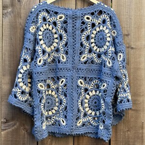 May include: A blue and cream crochet top with a floral pattern. The top is made of a soft yarn and has a loose, open design. The crochet work is intricate, with detailed floral motifs throughout the garment. The top is hanging on a wooden hanger.
