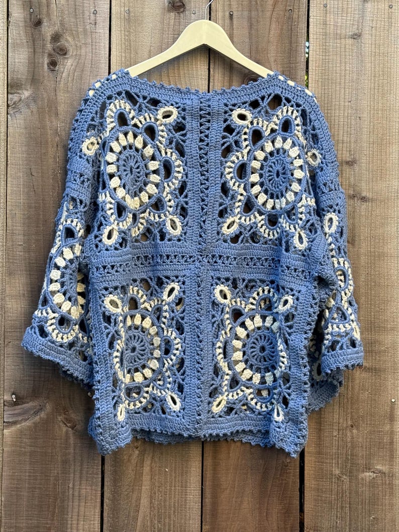 May include: A blue and cream crochet top with a floral pattern. The top is made of a soft yarn and has a loose, open design. The crochet work is intricate, with detailed floral motifs throughout the garment. The top is hanging on a wooden hanger.