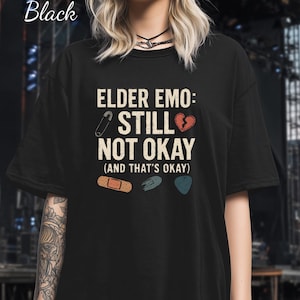 May include: Black t-shirt with the text "ELDER EMO: STILL NOT OKAY (AND THAT'S OKAY)". The design includes a safety pin, a broken heart, a bandage, and guitar picks. The word "Black" is written in cursive at the top left.