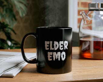 May include: A black ceramic mug with the words "ELDER EMO" in white, stylized text. Steam rises from the mug, suggesting a hot beverage. The mug sits on a wooden surface, with a newspaper and a glass teapot in the background.