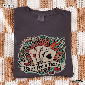 May include: Graphite-colored t-shirt with a graphic of four playing card aces, flames, and the text "She's From Texas." The design includes a decorative border with cherries and turquoise accents.