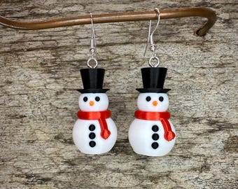 May include: A pair of snowman earrings with red scarves and black top hats. The earrings are made of white, red, and black polymer clay and have silver hooks.