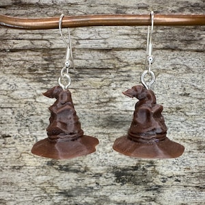 May include: A pair of brown Sorting Hat earrings. Each earring features a miniature, detailed replica of the iconic hat, complete with a pointed top and wide brim. The earrings hang from silver-colored fish hook ear wires.